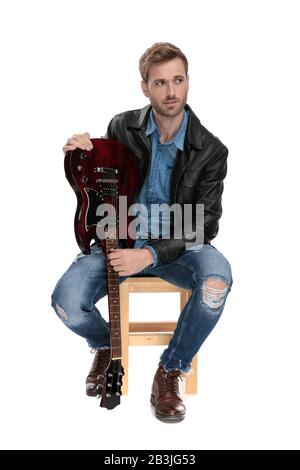 bel homme décontracté avec veste en cuir noir est assis sur une chaise en bois tenant sa guitare à l'envers et regardant sur le backroun de studio blanc Banque D'Images