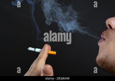 le jeune homme qui tient une cigarette dans ses mains et exhale la fumée sur un fond sombre. le préjudice causé par la nicotine et le tabac à proximité. Banque D'Images