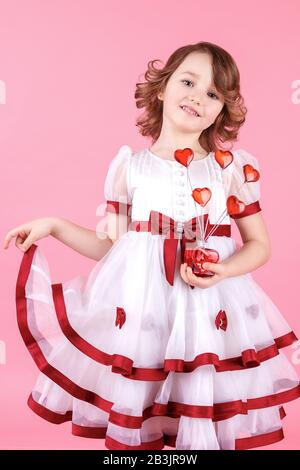 Portrait de la petite fille mignonne debout en robe blanche avec beignets ou coeur de verre sur le fond rose dans le studio. Concept d'amour et de soins Banque D'Images