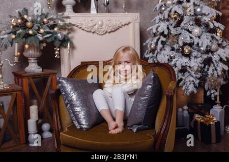 Un petit blond charmant est enveloppé dans un tapis dans une belle vieille chaise sous l'arbre de Noël. L'idée et le concept d'une bonne année et de Noël Banque D'Images