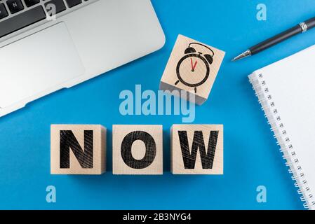 Word NOW et réveil sur des cubes en bois sur fond de bureau bleu avec bloc-notes et ordinateur portable Banque D'Images