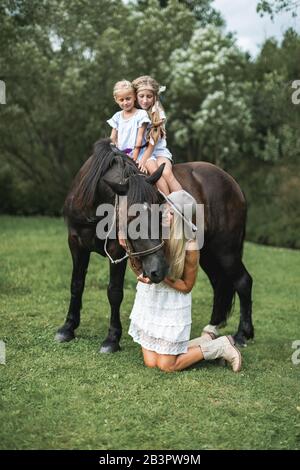 Jolie famille, mère et deux filles portant des vêtements style décontracté boho, marchant à l'extérieur sur le pré sur un cheval brun. Jolie jeune mère Banque D'Images