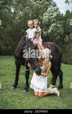 Jolie famille, mère et deux filles portant des vêtements style décontracté boho, marchant à l'extérieur sur le pré sur un cheval brun. Jolie jeune mère Banque D'Images