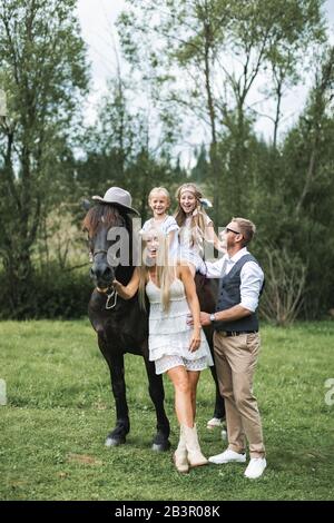 Famille heureuse, parents et enfants, profitant de la présence de cheval. Les filles sont à cheval, portant un chapeau. Homme et femme parents ayant plaisir et Banque D'Images