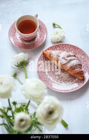 Une table du matin avec une tasse de thé avec croissant et des fleurs blanches de ranunculus floues Banque D'Images