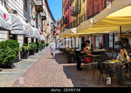 Les clients du restaurant en plein air se trouvent dans une rue pavée étroite, parmi des maisons colorées de la vieille ville d'Alba, en Italie. Banque D'Images