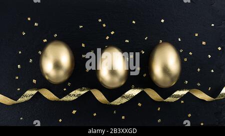 Œuf d'or avec des confettis décorés de brillance dorée, ruban pour bannière, carte de vœux, publicité sur les médias sociaux. Œufs d'or sur fond de pierre noire. Plat l Banque D'Images