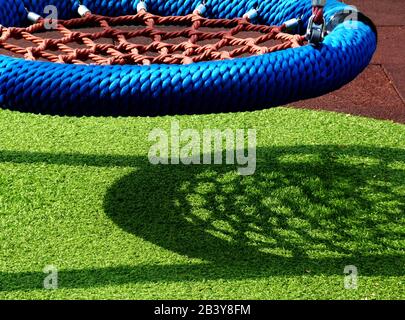 terrain de jeu en vinyle circulaire suspendu avec forte ombre sur l'herbe artificielle verte dans le parc public dans les couleurs bleu et rouge. fort soleil. Banque D'Images