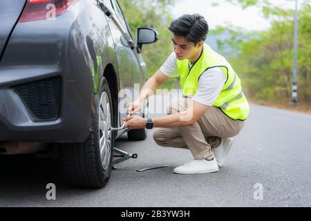 Jeune homme asiatique avec gilet de sécurité vert changeant le pneu crevé sur sa voiture en desserrant les écrous avec une clé de roue avant de soulever le véhicule Banque D'Images
