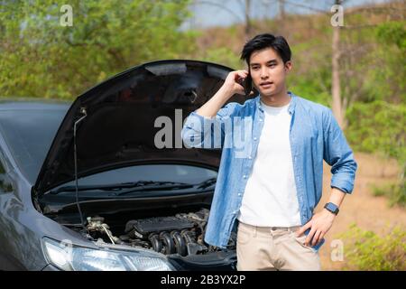 Jeune homme asiatique malcontent de parler sur un téléphone portable devant le capot ouvert cassé voiture Sur Country Road Téléphone Pour l'aide. Une Voiture Cassée Sur La Route I Banque D'Images