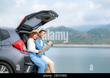 Jeune couple asiatique avec des jumelles homme-touriste tenant avec une petite amie souriante assis près de la carte sur le coffre de voiture et regardant l'appareil photo en rural f Banque D'Images