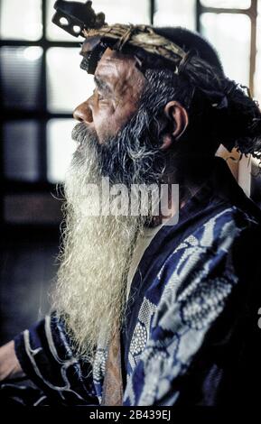 Un homme âgé d'Ainu avec une longue barbe et portant une couronne de fibres de bois pose dans la robe traditionnelle à l'intérieur de sa maison rurale sur l'île d'Hokkaido dans le nord du Japon. Ses vêtements ont des motifs pour se défaire des mauvais esprits. L'homme de 76 ans était l'un des seuls 300 Ainu pur-blooded (prononcé I-noo) vivant quand cette photographie historique a été prise en 1962. Depuis cette époque, les Ainu s'assimilent à la société japonaise et à leurs vieux métiers d'art, les coutumes et les cérémonies ne sont maintenues en vie que dans des villages spéciaux pour les touristes. L'Ainu a été officiellement reconnu comme peuple autochtone du Japon en 2008. Banque D'Images