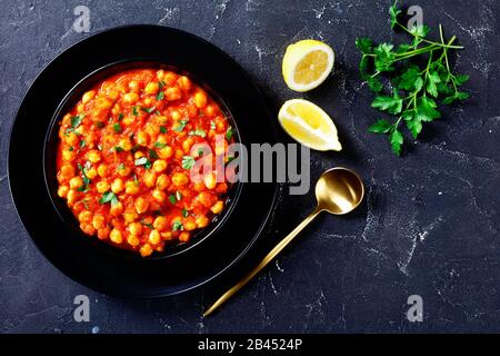 Plat de légumes indiens chana masala ou curry de pois chiches aux épices, sauce tomate saupoudrée de persil sur une plaque noire avec des quartiers de citron et une cuillère dorée, Banque D'Images
