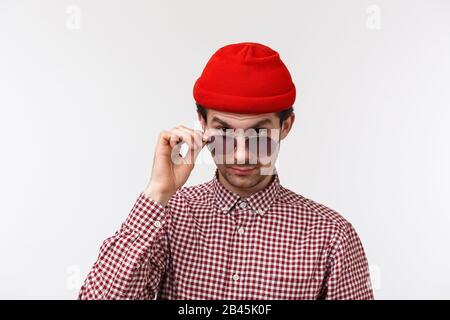 Sceptique et douteuse joli hipster jeune homme avec moustache, bonnet rouge, regarder de sous le front avec incrédulité, prendre des lunettes à faire Banque D'Images