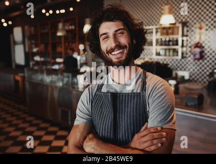 Portrait of smiling handsome young waiter standing at coffee shop Banque D'Images