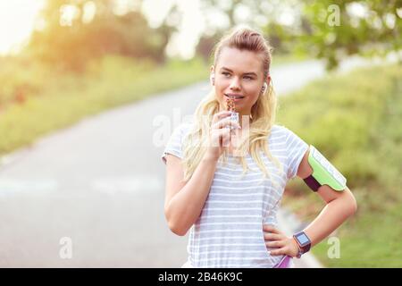 Une jeune femme moderne et sportive qui mangeait un bar d'énergie après une course dans le parc Banque D'Images