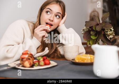 Image d'une jolie femme caucasienne portant des vêtements décontractés mangeant des fraises tout en prenant le petit déjeuner dans une chambre confortable Banque D'Images