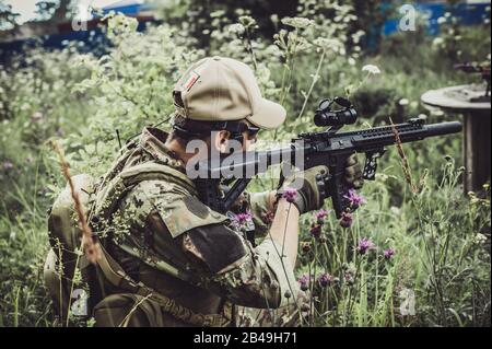 Un militaire ou un joueur aérodoux dans un costume de camouflage est assis dans l'herbe et vise d'un fusil automatique sur le côté Banque D'Images