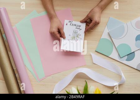 Horizontal de dessus plat composition de la fille mains tenant carte de vacances, bande de tulipes, papier d'emballage et ruban sur table en bois Banque D'Images