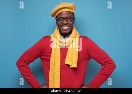 Bel homme noir afro-américain en pull rouge, chapeau jaune et écharpe, lunettes souriant regardant l'appareil photo en confiance. Émotion faciale positive. Banque D'Images