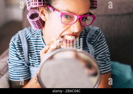 Jolie et belle jeune femme adulte à la maison, qui vérifie les rides de la peau ou les imperfections de l'âge devant un miroir - les boucles sur les cheveux et les lunettes pour l'âge adulte - les personnes actives et modernes vivent en bonne santé avec le stress Banque D'Images