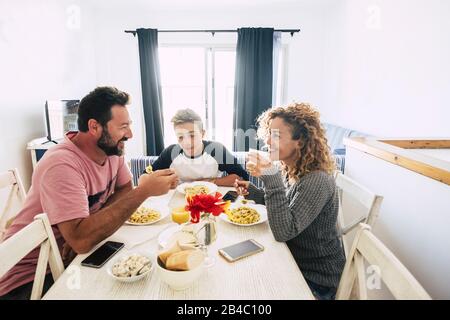 Les personnes gaies souriant en famille heureuse ont déjeuner ensemble à la maison - backgorund blanc et père de mère et fils d'adolescent mangeant des pâtes italiennes profitant de la vie ensemble Banque D'Images