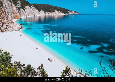 Lagon idyllique ensoleillé de la plage de Fteri avec littoral rocheux calcaire, Kefalonia, Grèce. Les touristes se détendent sous un parapluie près de l'eau de mer bleu émeraude turquoise claire avec un motif sombre au fond. Banque D'Images