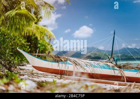 Gros plan de bateau banca local en bois devant la plage tropicale couverte de feuilles de palmiers sèches, paysage pittoresque à la lumière du soleil chaude, Palawan, Philippines. Banque D'Images