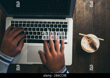 Vue de dessus de la station de travail avec ordinateur portable de technologie moderne et les mains pour l'utiliser et écrire - café ou cappuccino à son côté sur la table en bois - les gens au travail concept Banque D'Images