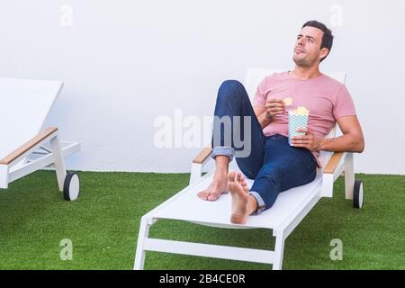 Beau homme facile mangeant la jonque fod à la maison se trouve dans le coucher de soleil - profiter de l'activité de détente avec l'herbe artificielle de pré et le blanc minimal backgorund Banque D'Images