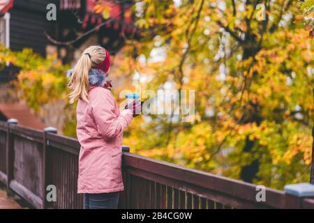 Une femme blonde adulte tenant une tasse dans ses mains se tient sur un pont en bois et regarde dans une forêt d'automne Banque D'Images