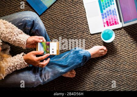 Une femme adulte à la mode est assise sur le sol et travaille à discuter avec des appareils technologiques comme un téléphone et un ordinateur portable - une fille non reconnaissable utilise les médias sociaux à la maison - les gens modernes avec connexion Internet Banque D'Images