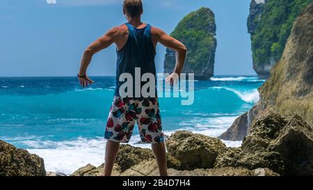 Homme devant les vagues de l'océan dans la Côte de Tembeling sur l'île de Nusa Penida, Immense Rocher en arrière-plan. Bali Indonésie. Banque D'Images