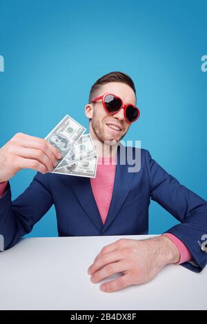 Jeune homme chic et riche en veste bleue et lunettes de soleil glamour, qui détient trois billets de banque en dollars tout en étant assis au bureau Banque D'Images