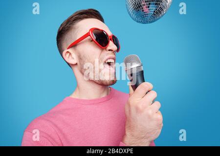 Jeune homme glamour dans des lunettes de soleil en forme de coeur et pullover rose tenant le microphone par sa bouche tout en chantant isolément Banque D'Images