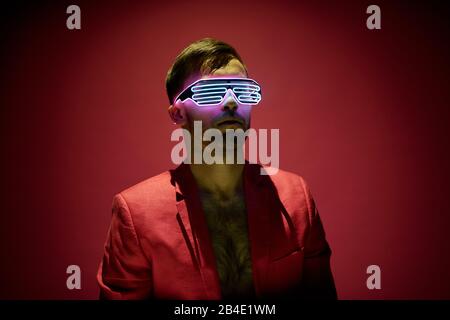 Jeune homme contemporain dans une veste rouge et des lunettes de haute technologie debout dans l'obscurité contre fond de marron en isolement Banque D'Images