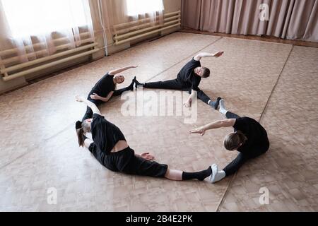 Quatre jeunes danseurs flexibles dans l'avtivewear assis avec des jambes tendues pendant l'entraînement sur le sol du studio de danse moderne Banque D'Images