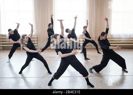 Trois filles et quatre gars en activewear noire debout sur le sol avec des jambes tendues et des genoux pliés tout en faisant de l'exercice dans le studio de danse Banque D'Images