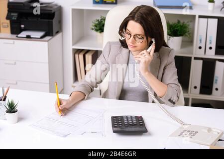 Jolie jeune femme d'affaires élégante assise au bureau, consultant le client au téléphone et prenant des notes dans le document Banque D'Images