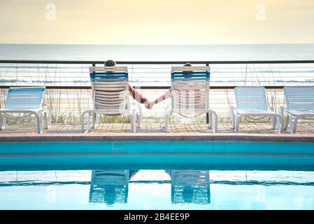 USA, Folrida, Daytona Beach, couple dans des chaises de plage près de la piscine, tenant les mains, surplombant la mer Banque D'Images