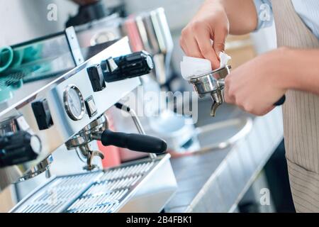 Vue rognée du jeune barista adulte dans un uniforme de tablier travaillant au restaurant, filtre de nettoyage dans une machine à café professionnelle, à l'aide d'une serviette Banque D'Images