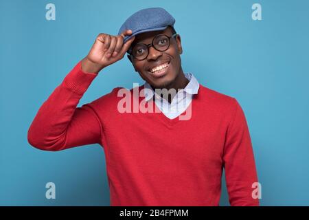 Bel homme noir afro-américain en sweat rouge, chapeau bleu, lunettes souriant regardant l'appareil photo confiant. Émotion faciale positive. Banque D'Images