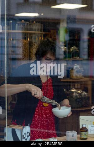 Blick durch eine Schaufensterscheibe auf ein Frau. Sie füllt Getreide in eine weiße Schüssel, Detail aus einem Unverpacktladen. Banque D'Images