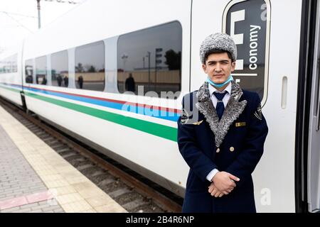 Chef d'orchestre dans un chapeau à fourrure traditionnel et masque facial contre Coronavirus devant le train rapide, la gare de Samarqand, Samarkand, Ouzbékistan Banque D'Images