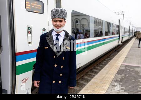 Chef d'orchestre dans un chapeau à fourrure traditionnel et masque facial contre Coronavirus devant le train rapide, la gare de Samarqand, Samarkand, Ouzbékistan Banque D'Images