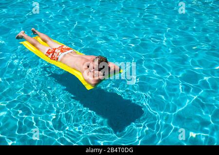 Homme imreconnaissable dans un short de bain coloré flottant sur un lilo gonflable sur une piscine bleu vif Banque D'Images