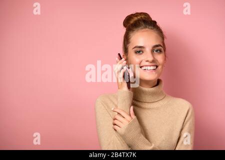 Une fille avec une peau propre et un grand pain se tient sur un arrière-plan rose souriant, parle au téléphone et regarde dans le cadre. Banque D'Images