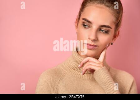 Une fille avec une peau propre et un haut pain se tient sur le fond rose et regarde pensivement sur le côté, propulsé sa joue avec le doigt Banque D'Images