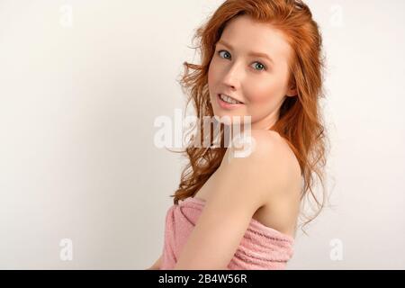 Une fille à poil rouge dans une serviette rose se tient sur un fond blanc à demi-tour avec un coeur peint avec de la crème sur l'épaule Banque D'Images