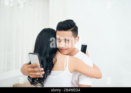 Jeune couple dans la chambre à coucher. L'homme infidèle sourire, c'est tricher et amant de SMS au téléphone tout en serrant sa petite amie Banque D'Images